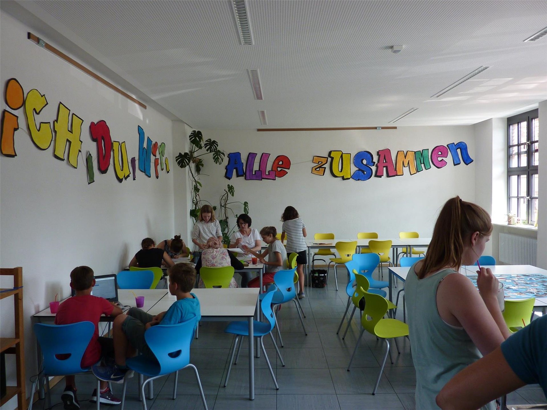 Mehrere Kinder und Jugendliche in einem Raum mit Tischen, bunten Stühlen und einem bunten Schriftzug an der Wand.